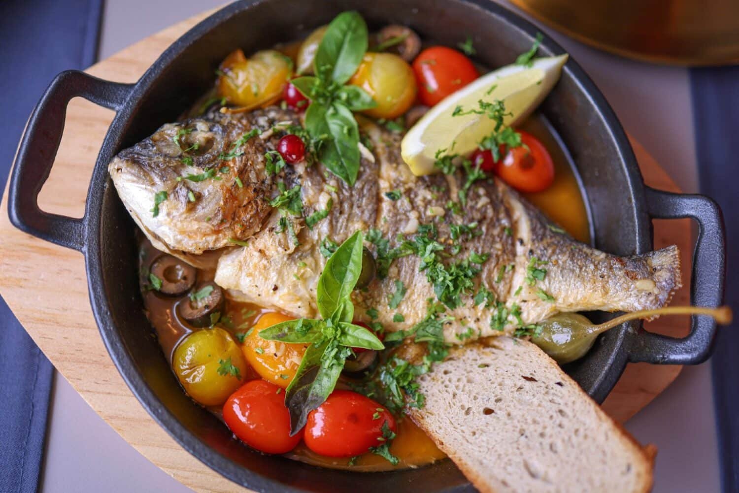 roasted fish with tomatoes and olives served with bread, fresh herbs and lemon slices.